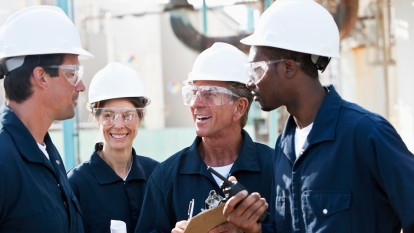 A group of four multiracial workers at a chemical plant talking. They are all in the same uniform, wearing white hardhats, safety glasses and navy blue coveralls. The oldest one is holding a clipboard. There are three men and one woman. The focus is on the people in the middle.