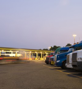 Number of semi trucks of various models and colors in the future on the night truck stop with a lit gas station and blurred lights of a passing semi truck, and a reflection of the lights on the chrome parts of this big rigs.