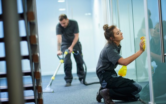 a female cleaning contractor is polishing the glass partition offices whilst In the background a male colleague steam cleans an office carpet in a empty office in between tenants.  .The female is smiling to camera.