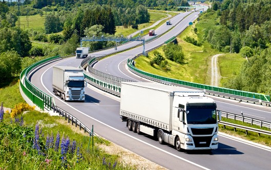 White trucks drive along an asphalt highway with electronic toll gate in a wooded landscape. View from above. Sunny summer day with blue skies and white clouds.