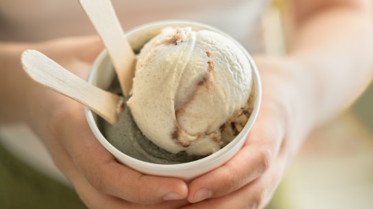 Closeup of organic, non-dairy, nut free, vegan ice cream held by a young woman. Made with coconut and sweet potato. Vanilla chocolate swirl and fennel licorice flavours, with 2 wooden spoons.