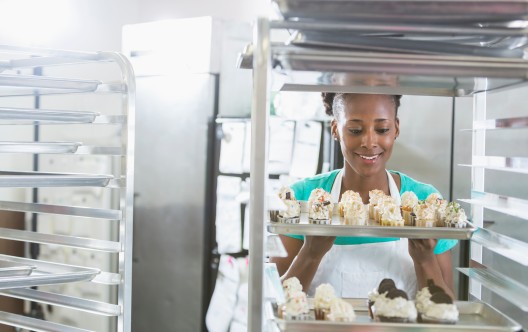 An African American woman wearing an apron, working in the commercial kitchen of a bakery. She is standing behind a rack of baked goodies. She is a successful minority small business owner.