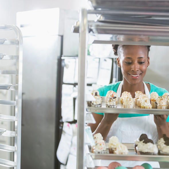 An African American woman wearing an apron, working in the commercial kitchen of a bakery. She is standing behind a rack of baked goodies. She is a successful minority small business owner.