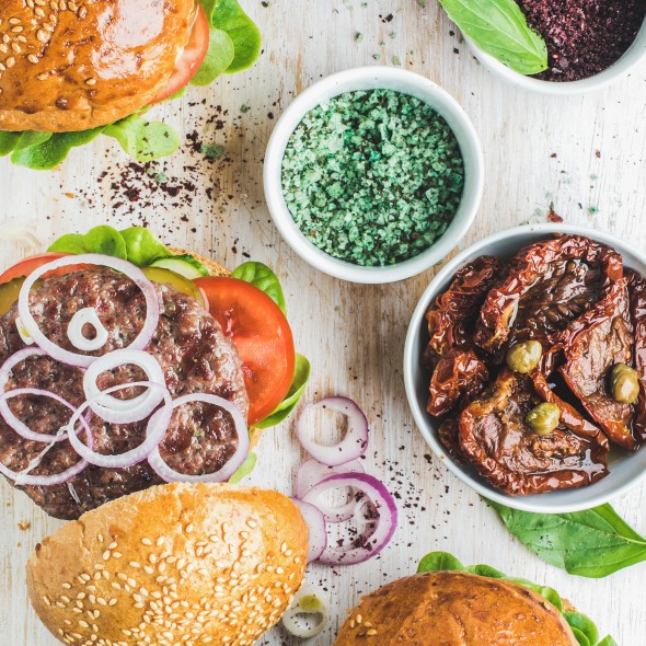Homemade beef burgers with onion, pickles, fresh vegetables, sun-dried tomatoes and tomato sauce on serving wooden board over blue painted background. Top view, vertical composition