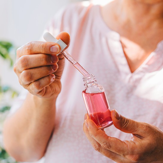 A close-up image of a mature woman holding a glass bottle with pink serum and a dropper. The photo highlights self-care and skincare routines in a naturally lit environment with green plants in the background, symbolizing health and well-being.
GettyImages-2185727212.jpg