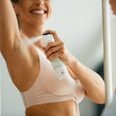 Close-up of happy woman spraying her underarm with deodorant while looking herself in a mirror.
GettyImages-1349701180.jpg
