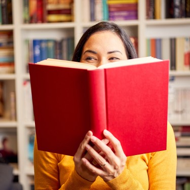 Smiling beautiful young female reading book standing against bookshelf at home
GettyImages-1381521896.jpg