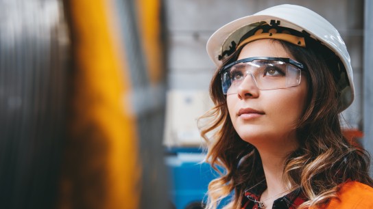 Portrait of young businesswoman with white helmet looking up and seen from the industrial steel cable reel for crane in factory warehouse. She is testing and working with winding wire and cable drum accessories metal wire spool reel wear resistance.