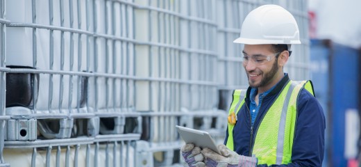 Young adult Hispanic man is chemical engineer checking inventory shipment of chemical crates on oil and gas pipeline job site. Man is wearing safety gear and using a digital tablet.