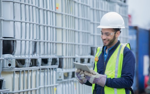 Young adult Hispanic man is chemical engineer checking inventory shipment of chemical crates on oil and gas pipeline job site. Man is wearing safety gear and using a digital tablet.