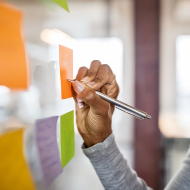 Close up of female hand writing suggestion on sticky note on glass wall. Business people brainstorming ideas in office.