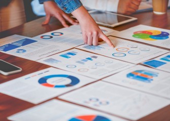 Paperwork and hands on a board room table at a business presentation or seminar. The documents have financial or marketing figures, graphs and charts on them. There is a digital tablet and laptop on the table