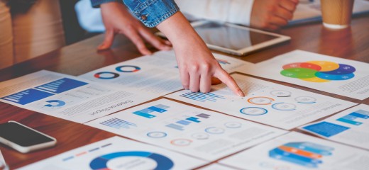 Paperwork and hands on a board room table at a business presentation or seminar. The documents have financial or marketing figures, graphs and charts on them. There is a digital tablet and laptop on the table