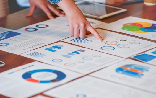 Paperwork and hands on a board room table at a business presentation or seminar. The documents have financial or marketing figures, graphs and charts on them. There is a digital tablet and laptop on the table