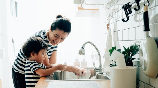 Asian mother with her baby boy washing dishes after cooking in kitchen at home.