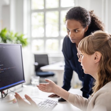 Businesswoman discussing with female coworker over coding on computer at desk in creative office