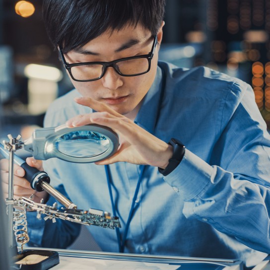 Close Up of a Professional Japanese Electronics Development Engineer in Blue Shirt Soldering a Circuit Board in a High Tech Research Laboratory with Modern Computer Equipment.
