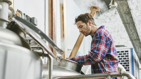 Low angle view of male craft beer worker in late 30s stirring batch in vat and doing quality check in Buenos Aires brewery.