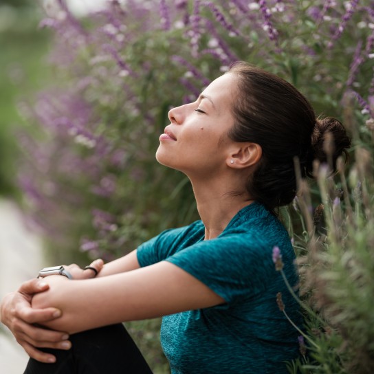 A latin woman sitting with her eyes closed and relaxing next to some flowers.