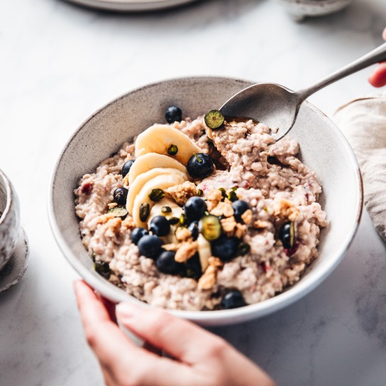 Close-up of a woman mixing oats flour, banana and blueberries in a bowl. Female making healthy breakfast in kitchen.