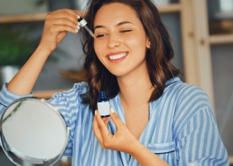 Young woman applying serum on her face while looking at mirror..