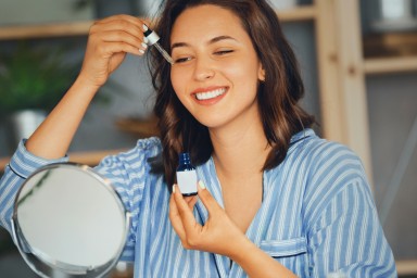 Young woman applying serum on her face while looking at mirror..