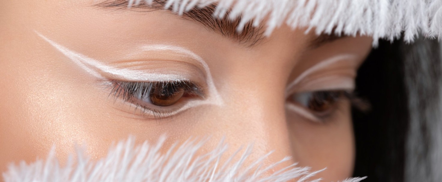 Portrait of a beautiful teenage girl with dark hair, with beautiful creative white makeup and healthy clean skin. She is holding a white peacock feather near her face. Makeup and cosmetology concept.