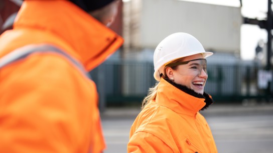 Female engineer smiling while talking with a worker at the shipyard. Two commercial dock workers in reflective uniform.