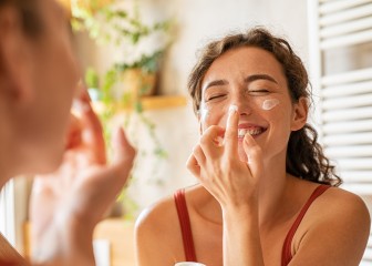 Playful young woman applying cream on nose. Cheerful girl holding green lotion jar standing in front of mirror applying moisturizer on nose. Beautiful woman taking care of skin by applying moisturizer every day in the morning.