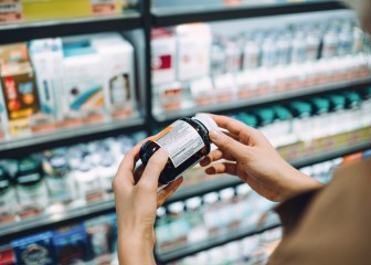 Over the shoulder view of young Asian woman browsing through medical products and reading the label on a bottle of medicine in front of the shelves in a pharmacy