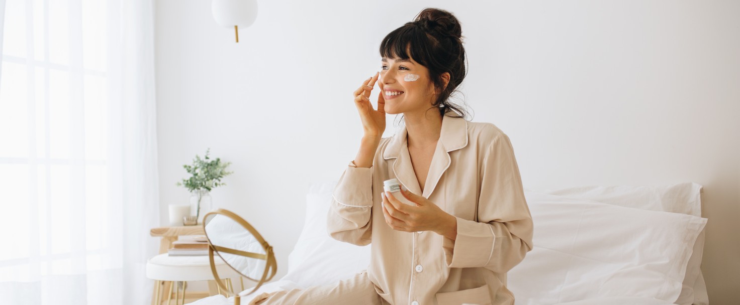 Happy woman doing routine skin care at home with beauty products. Woman sitting on bed at home and applying face cream.