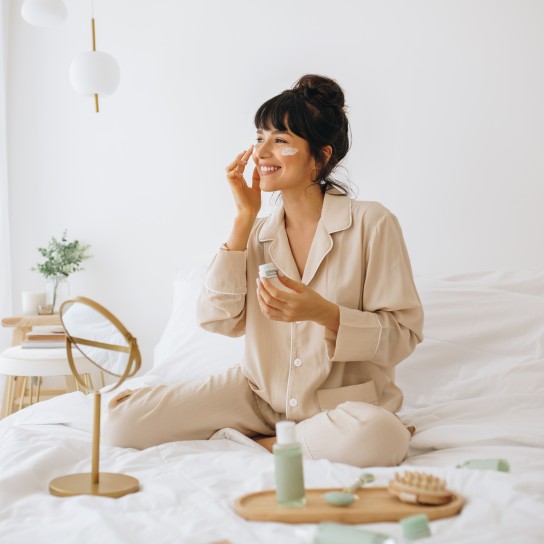Happy woman doing routine skin care at home with beauty products. Woman sitting on bed at home and applying face cream.