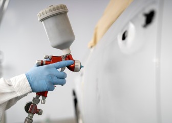 Close-up image of car painter with protective clothes and mask painting car using spray compressor in Auto Repair Shop.