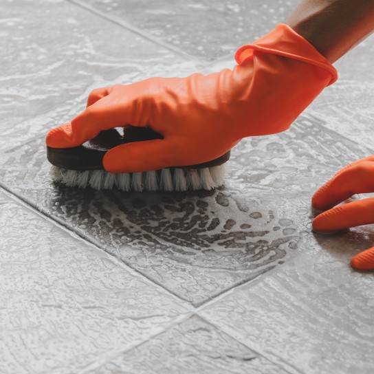 Hand of man wearing orange rubber gloves is use a hose to clean the tile floor.
