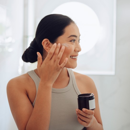 Beautiful and cheerful Asian woman standing in her bathroom, holding face cream and applying it to her cheeks and face.