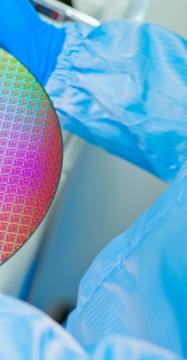 close up of asian male technician in sterile coverall holds wafer that reflects many different colors with gloves and check it at semiconductor manufacturing plant