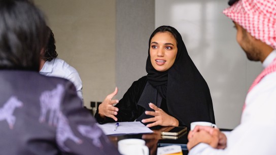 Waist-up view of Middle Eastern woman in traditional black abaya and hijab sitting at meeting table with development team and working out plans.