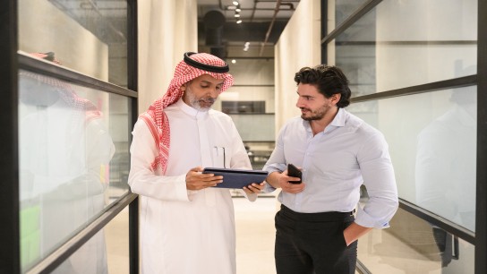 Front view of Middle Eastern men in western and traditional attire pausing to look at project data on digital tablet.