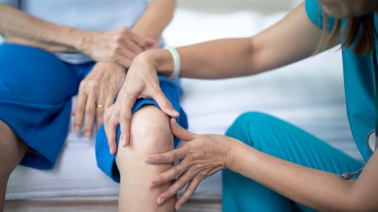 Female nurse home caregiver checking a knee to senior woman on bed at home.