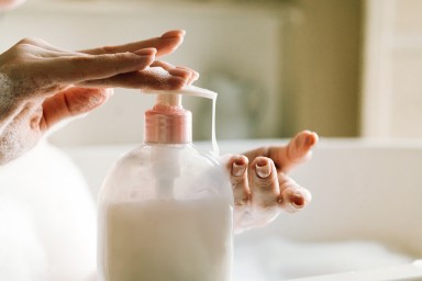 Dispenser with white liquid soap or shower gel, for intimate hygiene. Woman's hands pushing a button. White toilet soap in female hands against the background of a fragrant foam bath. Woman in white terry bathrobe washing her hands with soap. Relaxation and beauty treatments. Natural beauty, daily skincare routine. Moisturizing, cleansing