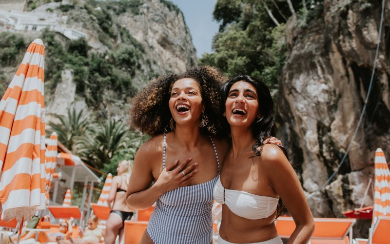 Two woman look delighted to be holidaying together in Italy. Views of Positano are visible behind them. They are surrounded by striped beach umbrellas and sun loungers. They wear stylish swimwear.