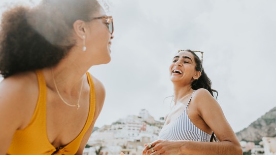 Two woman stand on a beach in Positano, Italy. They are wearing swimwear and sunglasses. They casually talk as they apply suncream in anticipation of a day of sunbathing at the beach. The woman in the foreground glances back at her friend in a bikini, who is the main focus of the image. They laugh.
