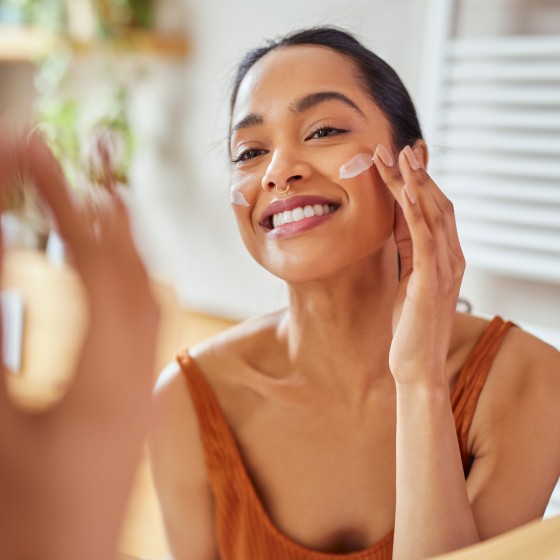 Portrait of smiling latin woman applying cream on her face while looking in mirror in her bathroom. Young multiethnic woman applying moisturizer on her cheek while looking in the mirror. Happy mexican girl using lotion every morning.