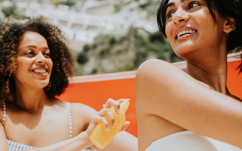 Two woman on sun loungers enjoy the beach on a hot day. One woman spritzes sun cream onto her friends back. They both look happy.