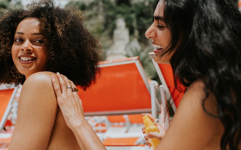Two woman on sun loungers enjoy the beach on a hot day. One woman spritzes sun cream onto her friends back. They both look happy.