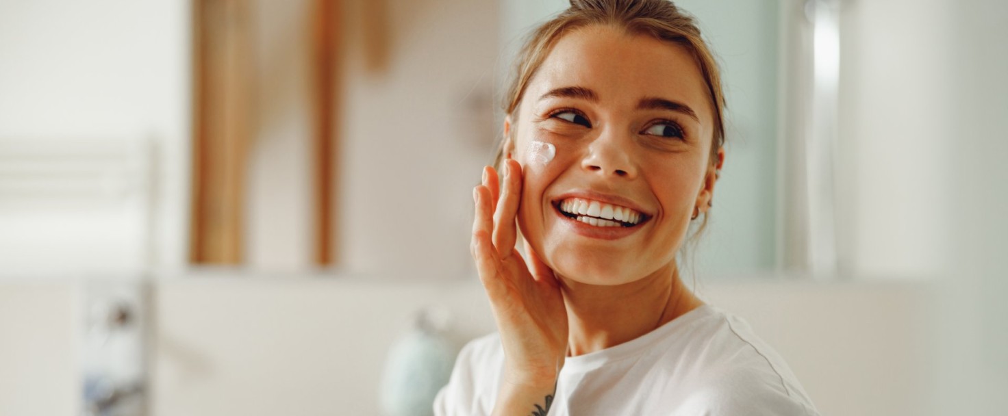 Happy woman applying hydrating moisturizer on her face sitting in bathroom. Home beauty routine