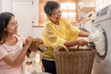 Attractive family loads laundry in washer appliance in kitchen at home. Asian senior elderly mom and daughter put dirty clothes to washing machine with her beagle dog. Domestic-House keeping concept.