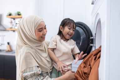 A mother in a hijab and her young daughter are putting a dirty cloth into a front-loading washing machine in a bright laundry room
