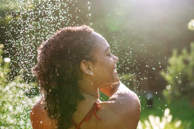 Rear view of beautiful young woman taking shower outdoors, in the middle of nature and lush foliage. Outdoor shower in garden.