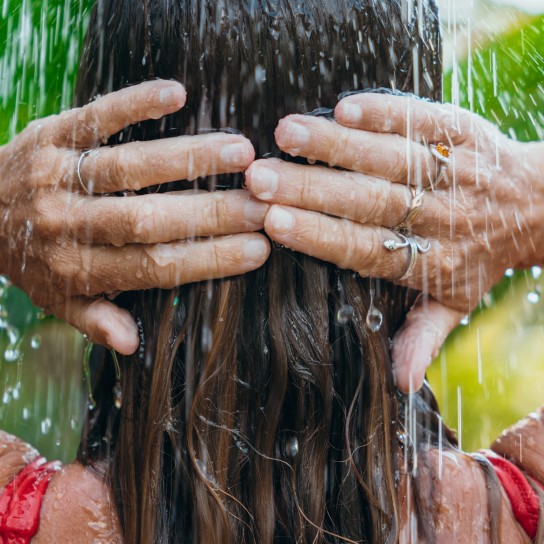 Woman with wet hair stands under a garden shower. Relaxing in a tropical setting, surrounded by lush greenery. Outdoor tranquility in a natural environment.